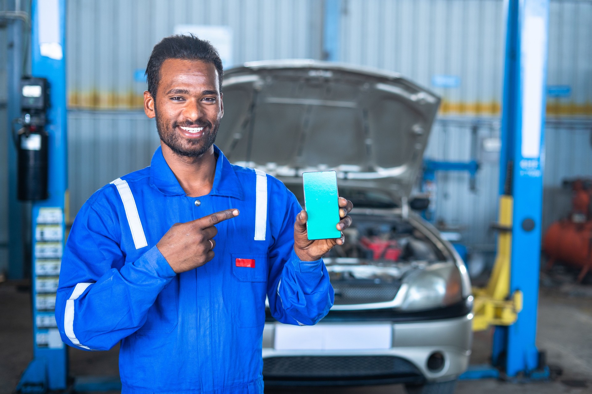 Smiling car mechanic showing green screen mobile phone with uniform in front of car by looking at camera - concept of online booking, advertisement and application promotion.